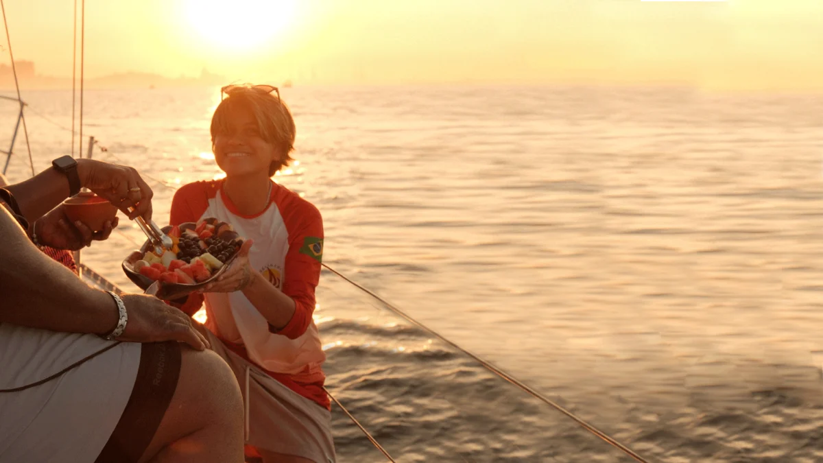membro da equipe da ddrio serve uma travessa de frutas tropicais cortadas para um passageiro no passeio de barco, iluminado pela luz dourada do fim de tarde ao fundo
