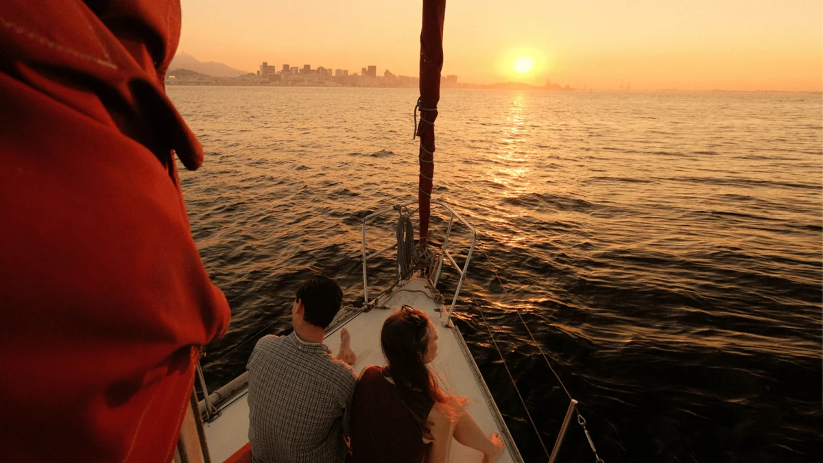 casal sentado na proa de um veleiro contempla o pôr do sol no passeio de barco
