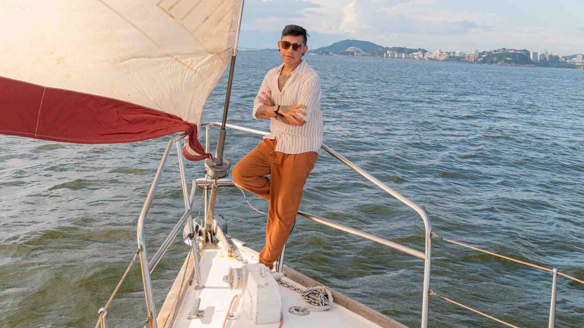 Um jovem de braços cruzados relaxa na proa de um veleiro, com o mar e uma cidade costeira ao fundo
