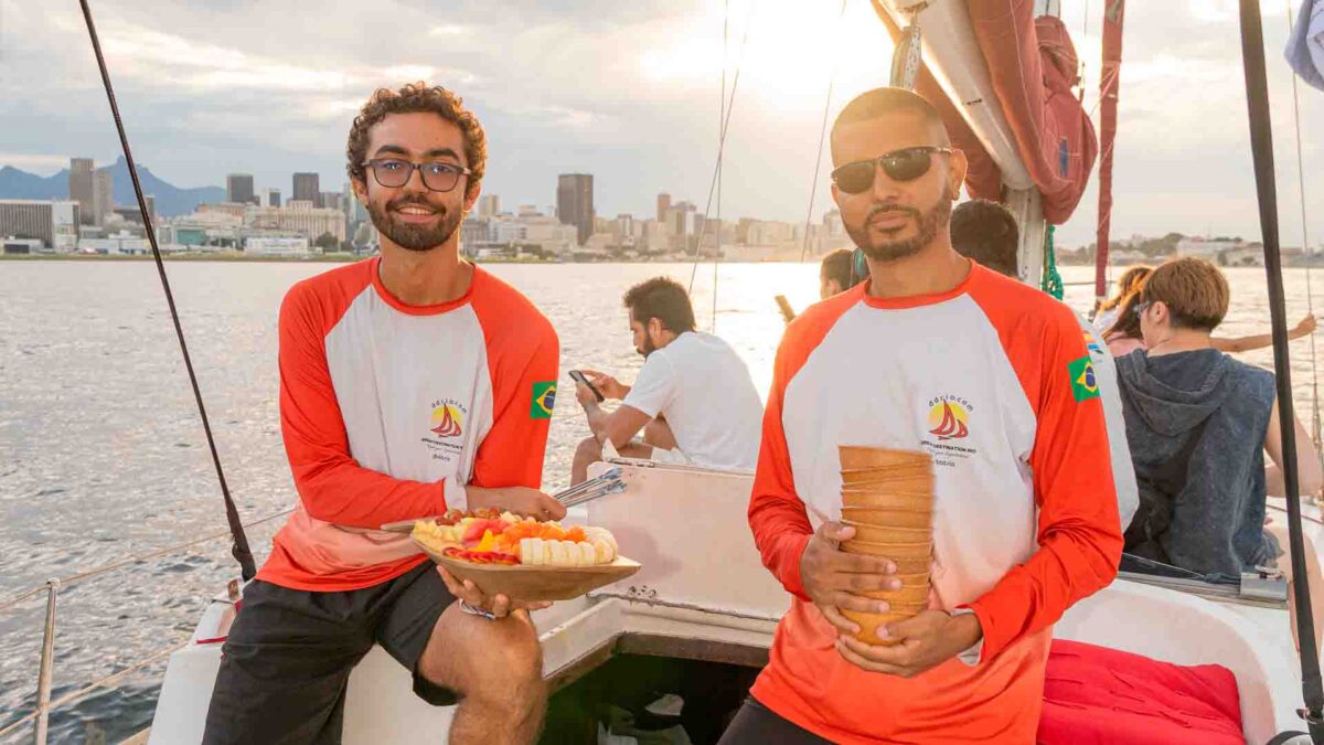 Dois tripulantes com camisas idênticas sorriem a bordo de um barco, segurando uma travessa de frutas cortadas e copos empilhados, com a paisagem urbana do Rio de Janeiro ao fundo