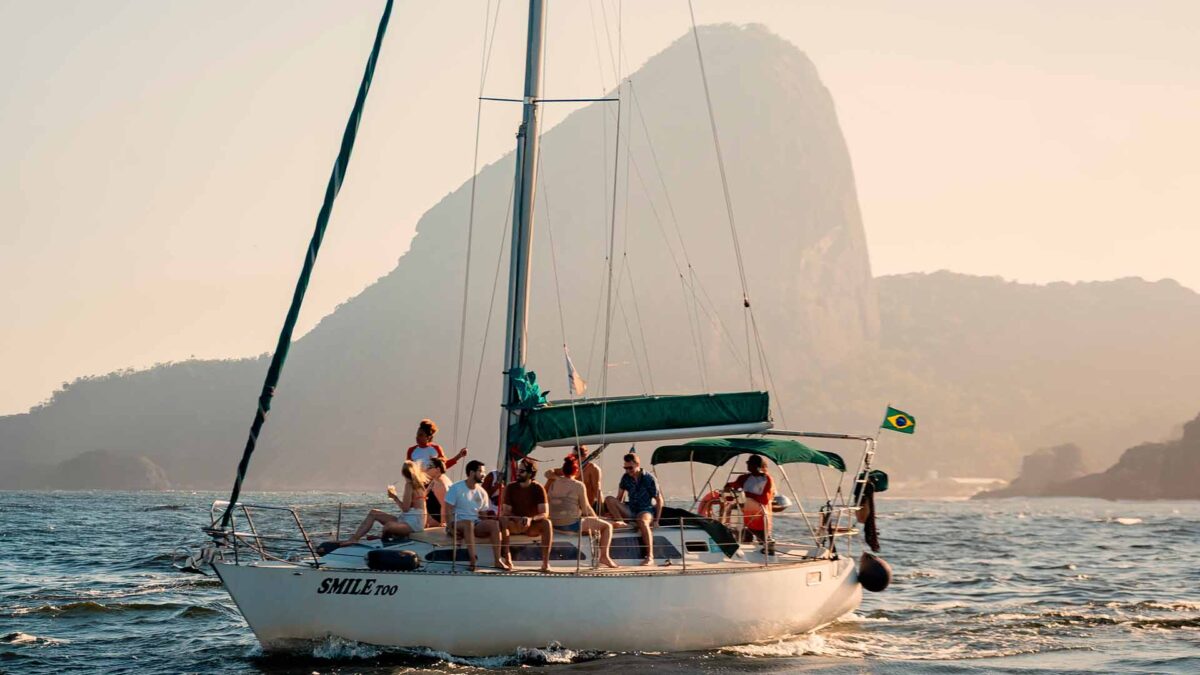 Um grupo de amigos navega num veleiro branco ao pôr do sol, com a silhueta do Pão de Açúcar ao fundo